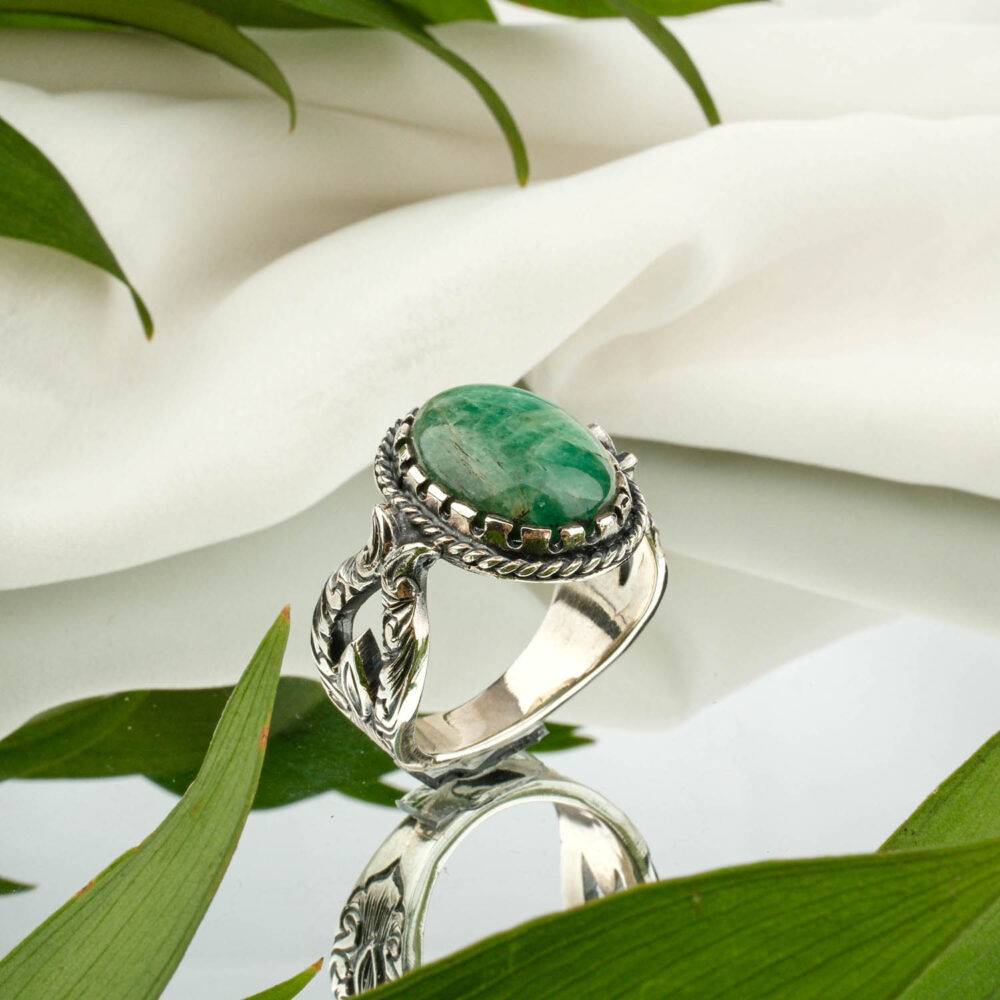 A faceted oval emerald men's ring in engraved sterling silver, on a reflective surface.