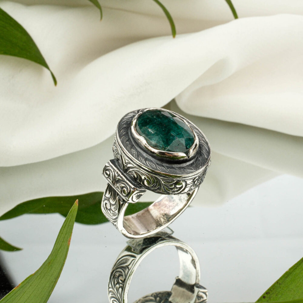 A faceted oval emerald men's ring in engraved sterling silver, on a reflective surface.