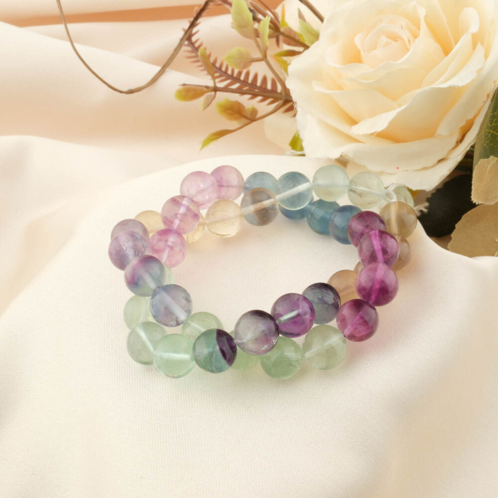 A 5A grade Rainbow Fluorite bracelet with 10mm polished beads, showing vibrant purple, green, and blue bands on a cream satin cloth.