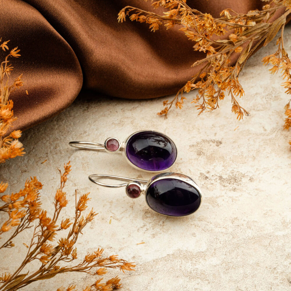 Premium Amethyst (oval cabochon) and Pink Tourmaline (round) two-stone earrings in 925 sterling silver, on a brown background.