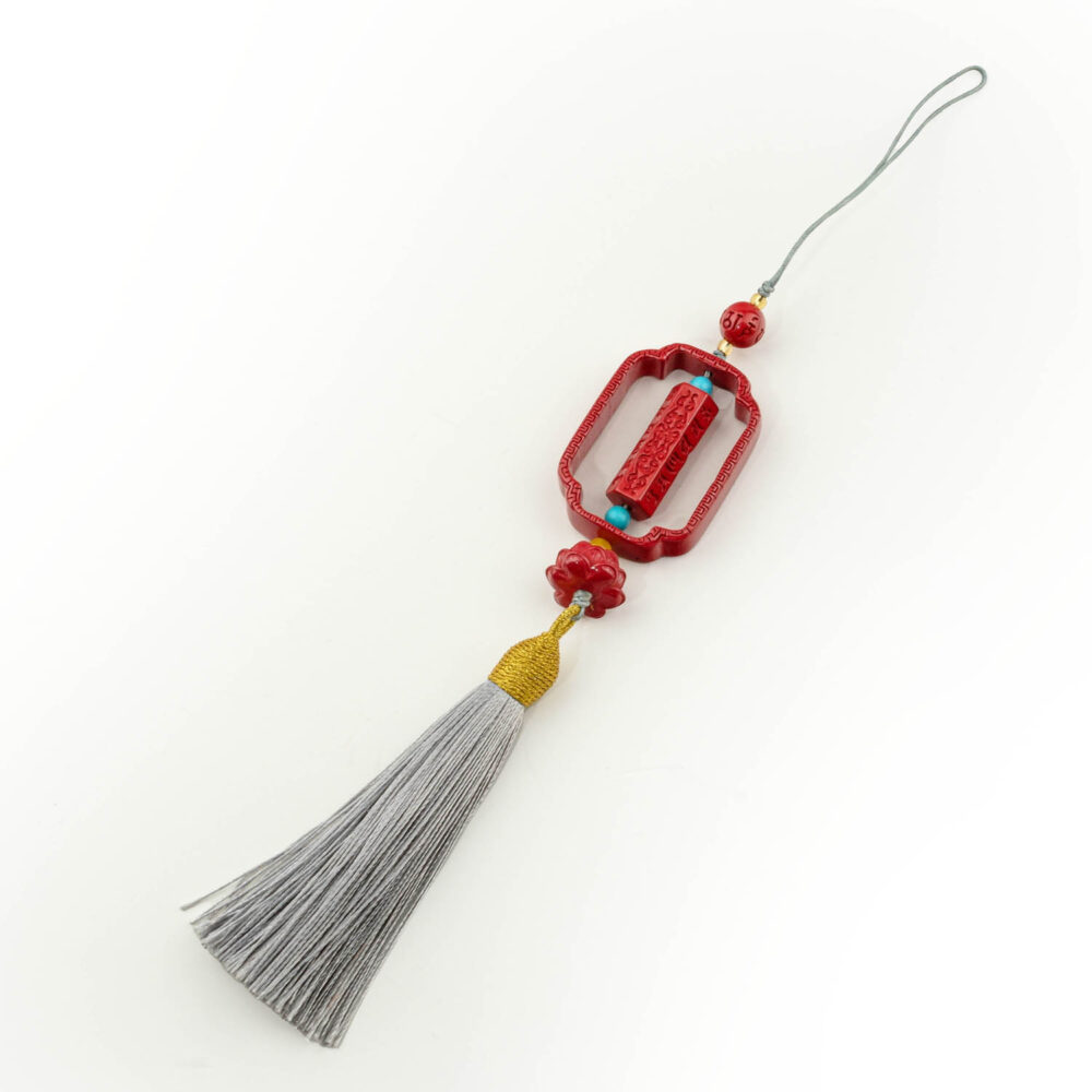 Amuleto de pergamino de cinabrio rojo tallado con borla plateada, sobre superficie blanca y madera.