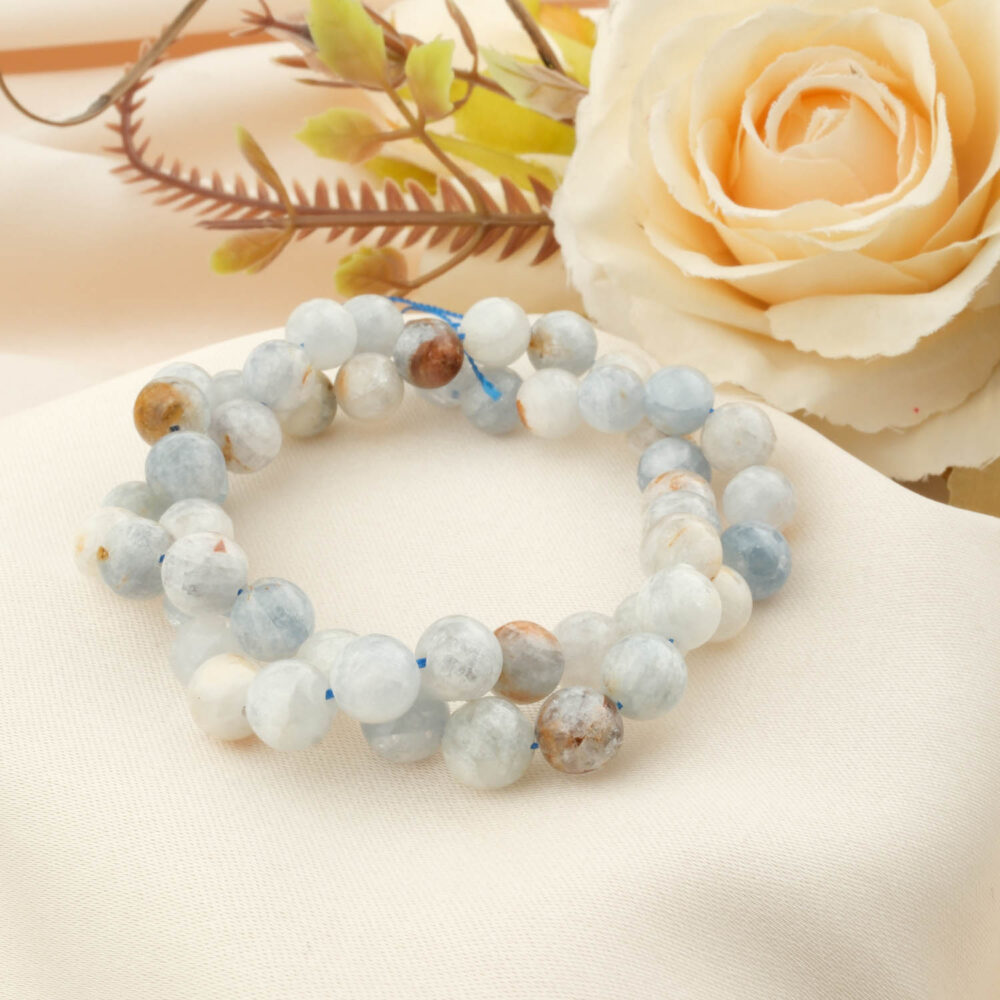 A strand of 5A grade 6mm round celestite beads with light blue, white, and brown hues, coiled on a cream silk background.