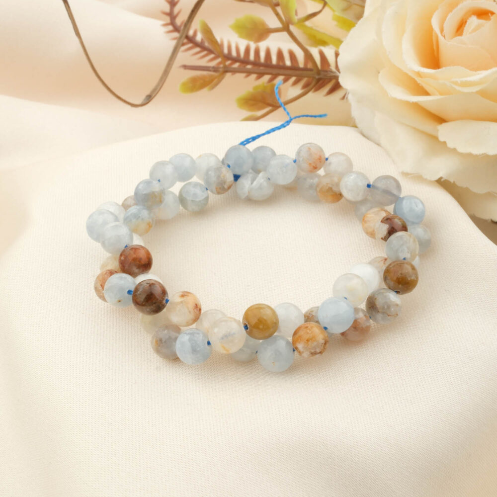 A strand of 5A grade 8mm round celestite beads with light blue, white, and brown hues, coiled on a cream silk background.