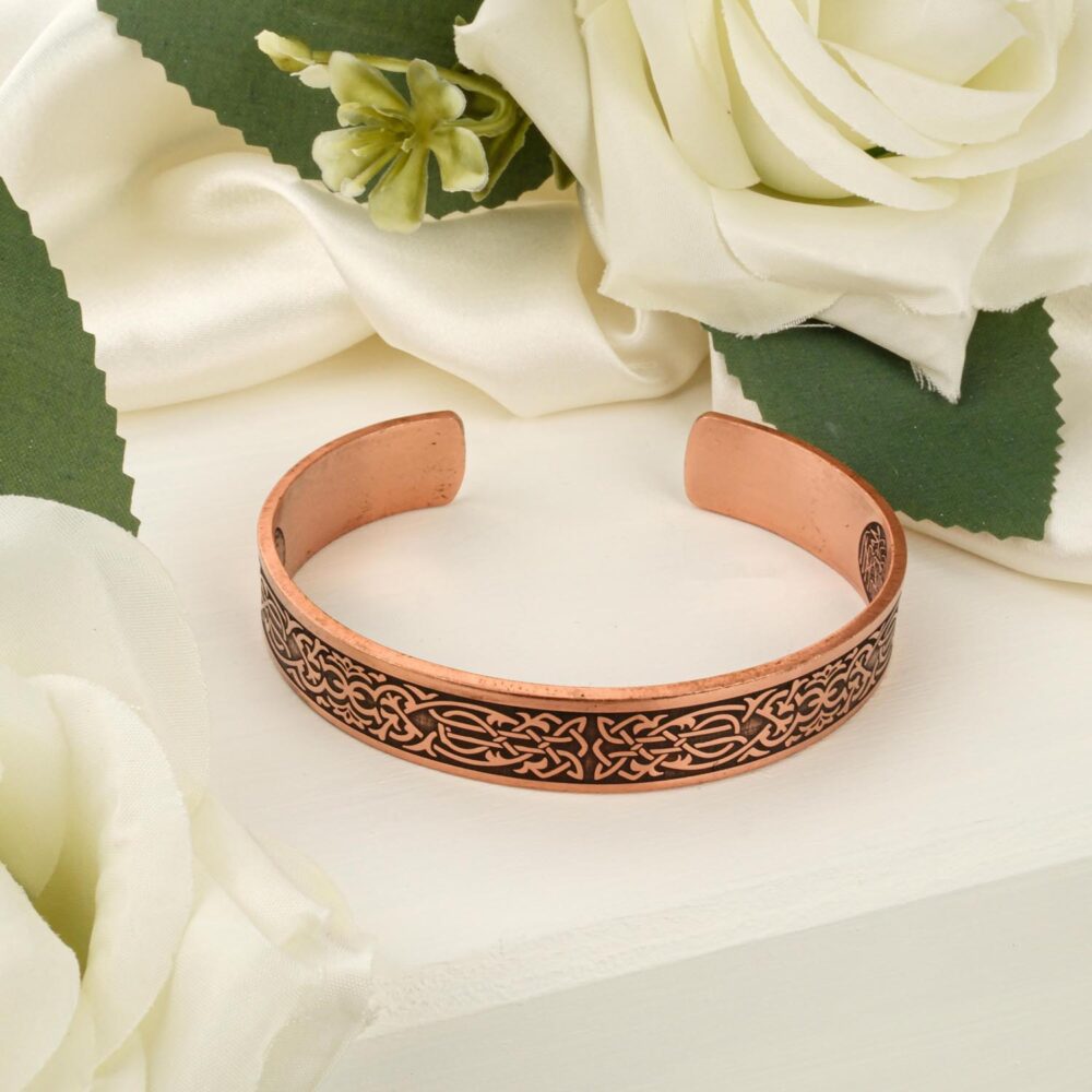 An engraved copper cuff bracelet featuring a detailed Celtic knot design, resting on a white textured surface with white roses.