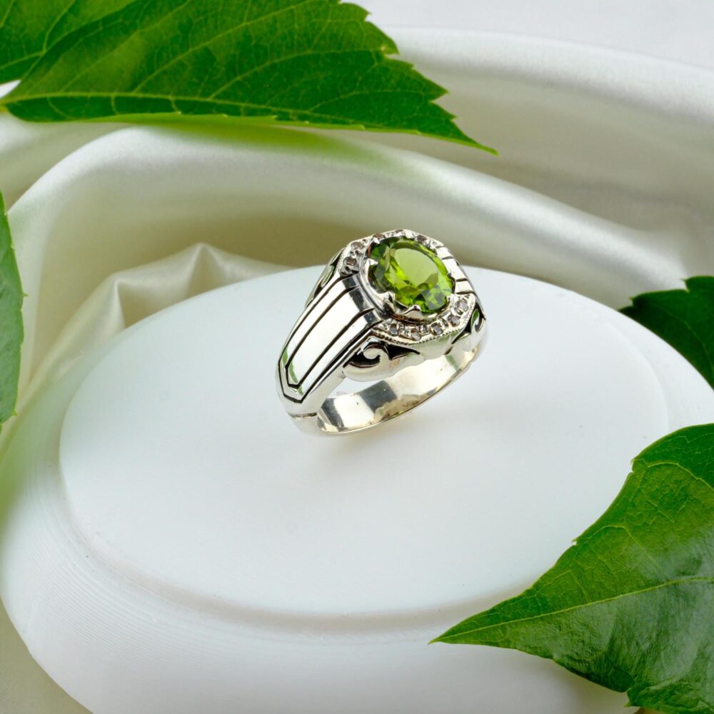 A men's sterling silver ring with a large oval faceted Peridot in a zircon halo, on a geometric engraved band, styled on a white surface.