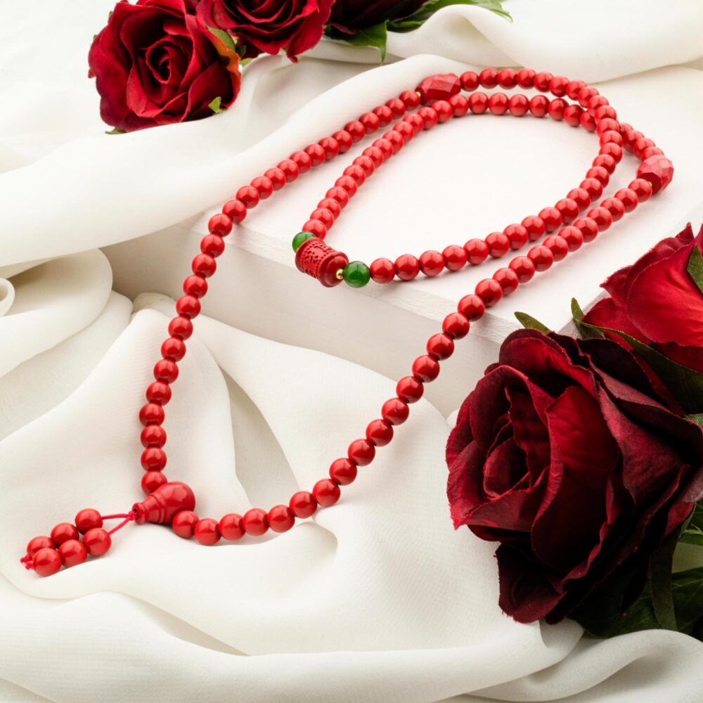 A traditional 108-bead Cinnabar mala necklace with a green accent stone, resting on white silk with red roses.
