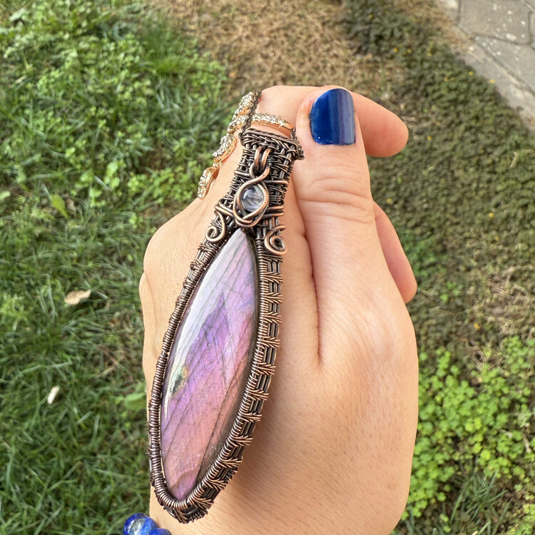 Eine handgefertigte Halskette aus Kupferdraht mit einem großen Labradorit-Stein mit violettem Schimmer, in einer Hand gehalten.