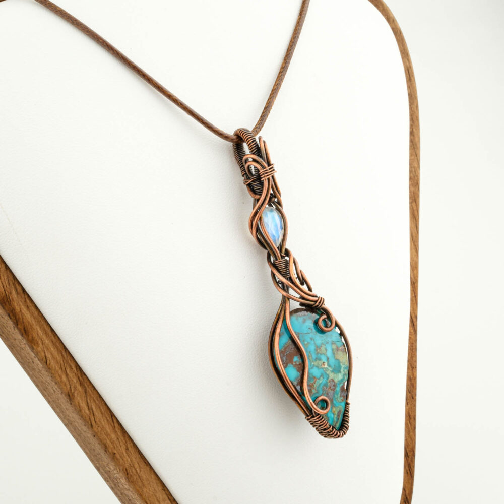 Colgante artesanal de cobre con gran piedra Turquesa ovalada y acento de Piedra Luna, sobre satén marrón.