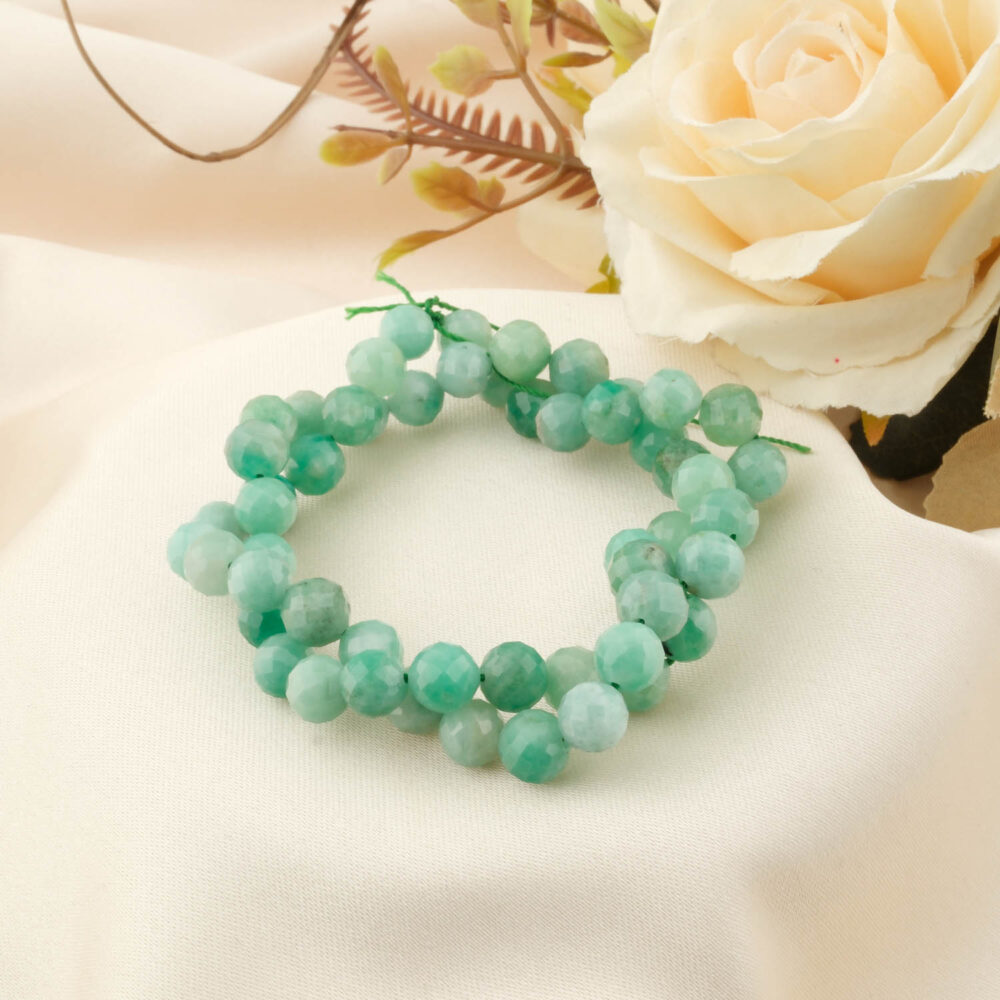 A strand of 5A grade 7-8mm faceted emerald beads with light green hues, coiled on a cream silk background with a white rose.