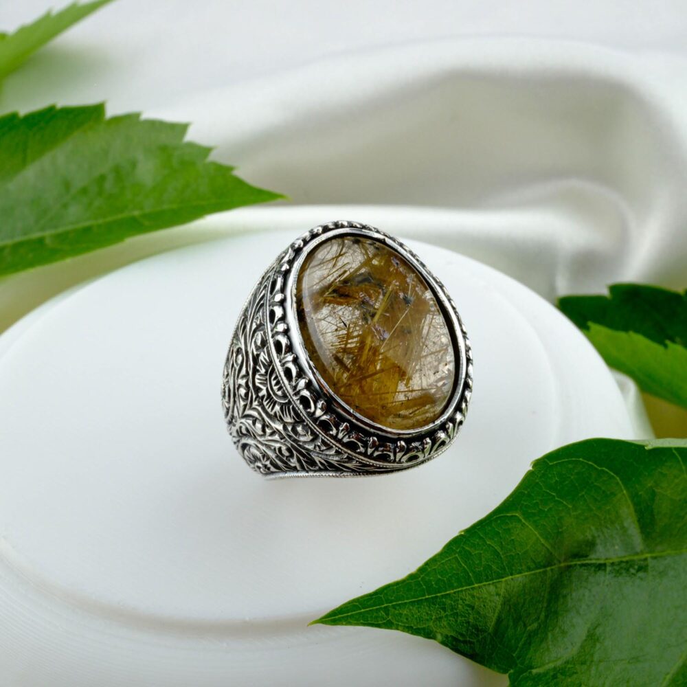 A large oval Golden Rutilated Quartz men's ring in an intricately engraved 925 sterling silver setting, on a white surface with green leaves.