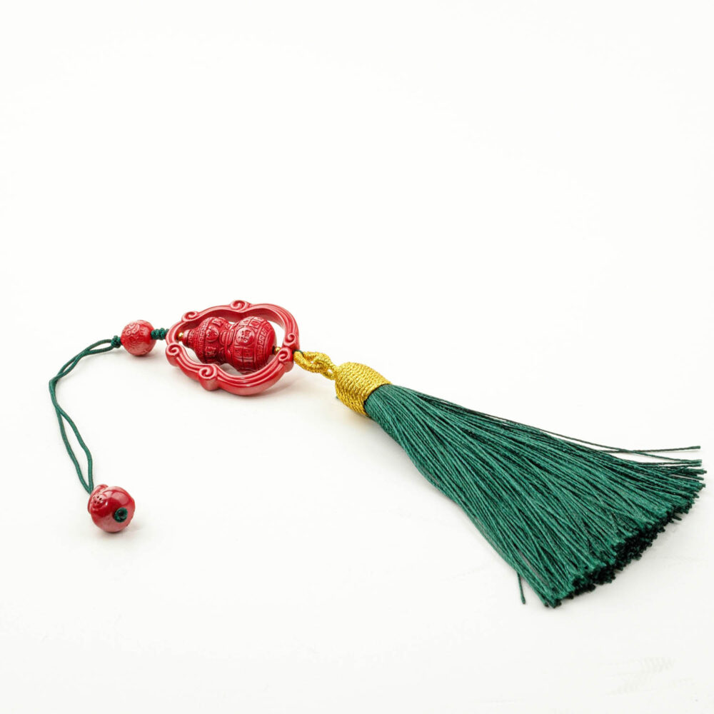 Amuleto Wu Lou de cinabrio rojo tallado con borla verde, sobre una superficie blanca y madera.