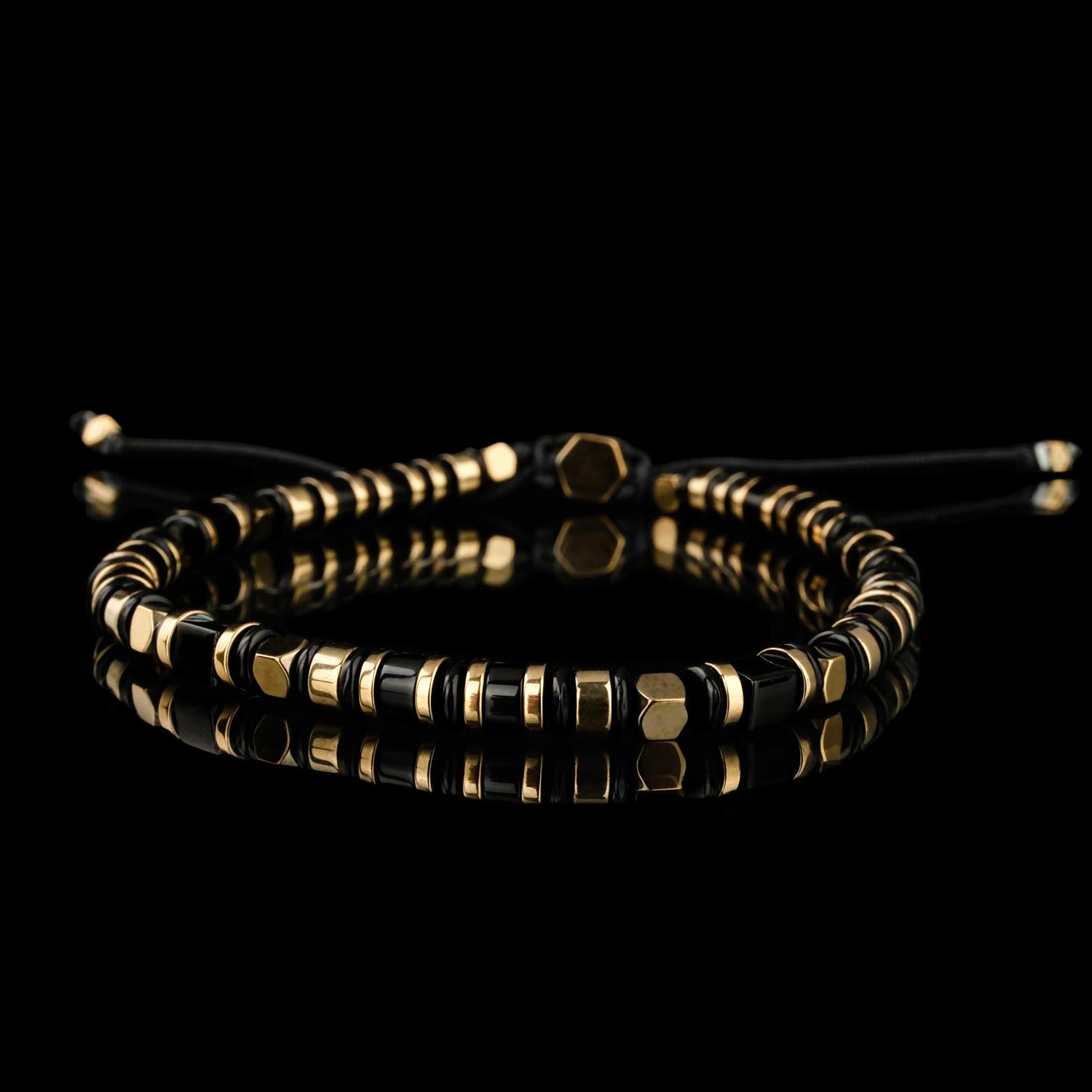 haematit-onyx-makramee-armband-goldton Ein elegantes Makramee-Armband mit schwarzen Onyx- und goldfarbenen Hämatit-Geometrieperlen an einer verstellbaren Kordel auf weißer Oberfläche.