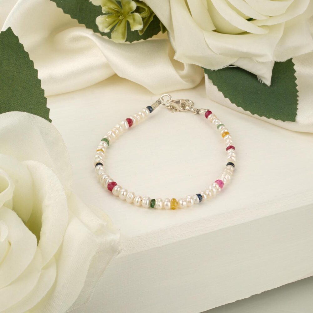 A delicate 3mm pearl bracelet with ruby, emerald, and sapphire accents, and a 925 sterling silver adjustable clasp, on a white display.