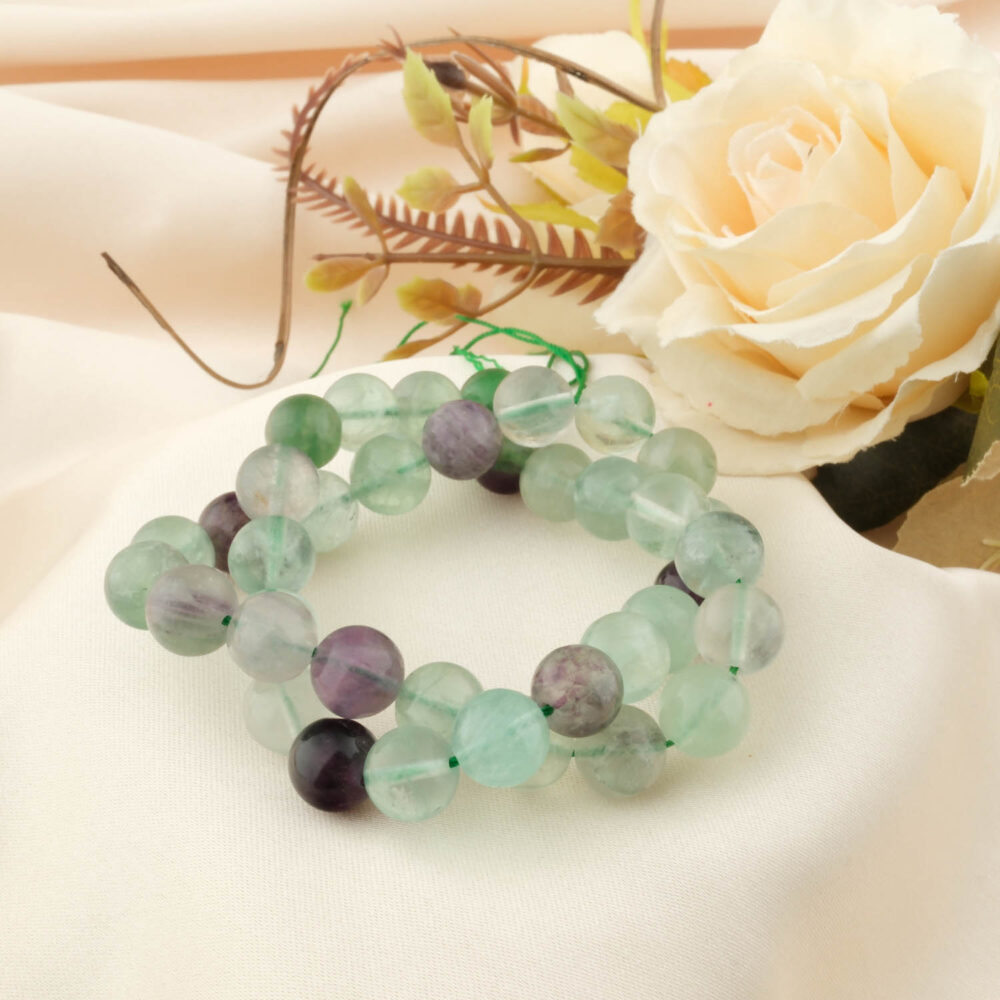 A natural Rainbow Fluorite bracelet with 10mm polished beads, showing green and purple bands, styled on a cream satin cloth with a rose.